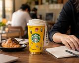 Starbucks cup with illustrations on a table with a croissant and notebook.